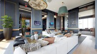 This stylish lounge space makes a striking first impression with its sophisticated design palette. The walls are finished in a deep slate blue, accented with gold trim that frames paneled sections, adding a sense of refined structure and visual depth throughout the room. The ceiling draws the eye upward, where two statement light fixtures anchor the space — a large medallion-style pendant with an ornate floral design and a bold teal drum shade pendant, both contributing dramatically to the room's upscale character.

The seating area is generously appointed with oversized white sofas and loveseats, layered with an eclectic mix of throw pillows in warm tones of copper, blush, teal, and taupe. Acrylic side tables and low-profile coffee tables provide functional surfaces while maintaining a light, modern aesthetic. Multiple flat-screen televisions are mounted on the walls, enhancing the space's utility as a social and entertainment hub.

Lush greenery, including a tall potted plant in a dark planter, introduces a natural element that softens the polished décor. Light wood flooring ties the design together, offering warmth against the cooler wall tones. Overall, the space exudes a contemporary yet welcoming ambiance ideal for social gatherings.