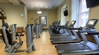 This fitness center combines classic architectural charm with modern exercise equipment to create a well-rounded workout environment. The space features warm hardwood flooring throughout, crown molding, and tall windows that allow natural light to fill the room, contributing to an inviting atmosphere. Pendant lighting hangs from the ceiling alongside recessed lights, providing ample illumination throughout the day and evening.

A row of Matrix treadmills and elliptical machines lines one side of the space, offering a comprehensive selection of cardio equipment for users of varying fitness levels. Strength training machines are positioned toward the rear of the room, visible beyond an arched passageway that adds a refined architectural touch and creates a sense of depth within the facility. A wall-mounted flat-screen television is positioned for easy viewing during workouts.

The facility's elongated layout allows for clear circulation between equipment, and a large mirror at the far end visually expands the space. The combination of neutral warm-toned walls, classical detailing, and contemporary fitness equipment gives this fitness center a polished and functional character.