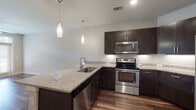 This single-floor residence showcases a contemporary design with a cohesive blend of modern finishes throughout. The kitchen serves as a central highlight, featuring dark espresso cabinetry paired with light granite countertops that extend across a peninsula with seating potential. A stainless steel appliance suite includes a French door refrigerator, electric range, built-in microwave, and dishwasher. Two sleek pendant lights hang above the peninsula, complementing the recessed lighting overhead. Rich hardwood-style flooring runs throughout the main living areas, adding warmth and continuity to the open layout.

The living area connects seamlessly to the kitchen, creating an open, functional flow. Bedrooms are finished with neutral-toned carpet and feature windows with blinds that allow natural light. The bathroom is well-designed with a navy blue vanity topped with granite, a framed mirror with wall sconces, wood-look flooring, and a tub-shower combination with tile surround. Neutral gray walls throughout the unit provide a clean, versatile backdrop that complements the modern aesthetic of the space.