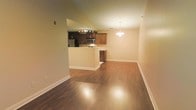 This single-floor space is designed with an open-concept layout that promotes a sense of openness and ease of movement throughout. The kitchen is a standout feature, showcasing rich dark wood cabinetry, stainless steel appliances including a microwave and refrigerator, and a smooth light-toned countertop that extends across a generous peninsula. The peninsula serves as a natural divider between the kitchen and the adjoining living and dining areas, making it ideal for entertaining or casual everyday use. Track lighting above the kitchen provides focused illumination, while a stylish chandelier anchors the dining area with a warm, inviting glow. The living and dining spaces are expansive, offering ample room for a variety of furniture arrangements. Warm-toned hardwood flooring runs throughout the entire space, adding continuity and a polished aesthetic. The neutral beige wall tones complement the flooring and cabinetry, creating a cohesive and welcoming atmosphere. White baseboards provide clean contrast and a classic finishing detail. Overall, the space balances functional design with an approachable, comfortable ambiance suited for both relaxed living and social gatherings.