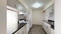 This single-floor unit offers a practical and efficiently designed layout across a modest footprint. The galley-style kitchen is a standout feature, equipped with ample white cabinetry on both sides, dark granite-style countertops, white appliances including a coil-top electric range and refrigerator, and a tile backsplash. Wood-look plank flooring runs throughout the kitchen and connecting hallways, lending a clean, cohesive aesthetic.

The layout includes two hallways that guide residents through the unit, connecting the kitchen and entry areas to the private spaces beyond. The spacious living area features neutral carpet and multiple windows fitted with blinds, allowing generous natural light to fill the room. A pendant light fixture adds a functional design accent overhead.

The bedroom is generously proportioned, featuring plush carpeting, a large window for natural light, and a built-in closet with a hanging rod for ample storage. The full bathroom features wood-look vinyl flooring, a single-sink vanity with white cabinetry, a tiled bathtub and shower combination, and a chrome towel bar. The overall palette of warm neutrals and white trim creates a clean, move-in-ready atmosphere throughout.