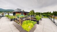 Located atop a modern building on Lonsdale Avenue in North Vancouver, this expansive rooftop amenity space offers a thoughtfully designed outdoor retreat with impressive panoramic views. The open-air terrace is defined by large-format porcelain tile paving throughout, providing a clean and contemporary aesthetic underfoot.

A standout feature of the rooftop is its extensive collection of raised garden planters, constructed from sleek dark metal and arranged across the terrace. These generously sized beds are actively cultivated with a variety of vegetables, herbs, and flowering plants, offering residents a true community gardening experience. Vertical support stakes within the planters indicate active growing seasons and a well-maintained, productive garden program.

A covered outdoor pavilion with warm wood-clad ceilings and a contemporary grey metal roof provides a sheltered gathering area. Integrated within this structure is a dedicated BBQ station, making it well-suited for outdoor cooking and entertaining. Adjacent seating areas invite residents to dine and socialize in a comfortable, partially shaded environment.

A lush artificial lawn area with lounge chairs faces outward toward the glass railing perimeter, where unobstructed views of the North Vancouver treetops, the city of Vancouver, Burrard Inlet, and the surrounding mountain range create a striking natural backdrop. The combination of greenery, functional amenities, and exceptional views makes this rooftop an exceptional communal outdoor space.