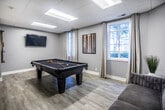This single-story community room in Memphis, Tennessee offers a well-appointed and versatile gathering space designed with modern aesthetics and practical amenities. Throughout the space, light wood-look flooring, neutral gray walls, and a drop ceiling with recessed panel lighting establish a clean, contemporary atmosphere.

The open-plan kitchen and dining area serves as the social heart of the room. The kitchen is outfitted with dark navy cabinetry, a herringbone-tile backsplash, stainless steel appliances, and a center island with a white quartz countertop and black bar stools. A striking brass multi-bulb pendant chandelier illuminates the space, while a window with sheer drapery brings in natural light. A round white dining table with gold-accented chairs provides a stylish spot for communal meals.

Adjacent to the kitchen, a lounge area features a textured accent wall in a bold geometric pattern, a white credenza, and table lamps that add warmth. Structural columns define the zones without fully enclosing them, maintaining an open, connected feel throughout.

A dedicated game room offers a full-size pool table, a wall-mounted television, a cue rack, and natural light from a curtained window, making it an ideal space for recreation and entertainment.