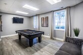 This single-story community room in Memphis, Tennessee offers a well-appointed and versatile gathering space designed with modern aesthetics and practical amenities. Throughout the space, light wood-look flooring, neutral gray walls, and a drop ceiling with recessed panel lighting establish a clean, contemporary atmosphere.

The open-plan kitchen and dining area serves as the social heart of the room. The kitchen is outfitted with dark navy cabinetry, a herringbone-tile backsplash, stainless steel appliances, and a center island with a white quartz countertop and black bar stools. A striking brass multi-bulb pendant chandelier illuminates the space, while a window with sheer drapery brings in natural light. A round white dining table with gold-accented chairs provides a stylish spot for communal meals.

Adjacent to the kitchen, a lounge area features a textured accent wall in a bold geometric pattern, a white credenza, and table lamps that add warmth. Structural columns define the zones without fully enclosing them, maintaining an open, connected feel throughout.

A dedicated game room offers a full-size pool table, a wall-mounted television, a cue rack, and natural light from a curtained window, making it an ideal space for recreation and entertainment.