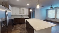 This contemporary single-floor residence showcases a thoughtfully designed open-concept layout with modern finishes throughout. The heart of the home is the chef-inspired kitchen, which features a striking two-tone design pairing rich dark wood lower cabinetry with crisp white upper cabinets accented by frosted glass panels. A generous quartz-topped island anchors the space and is illuminated by stylish pendant lighting. Stainless steel appliances — including a side-by-side refrigerator with ice maker, range with over-the-range microwave, and dishwasher — complete the culinary space. A classic subway tile backsplash adds a clean, polished touch. Warm hardwood-style flooring flows throughout the main living areas, complemented by neutral walls that create a bright and airy atmosphere. The living area features a ceiling fan and natural light from large windows. The bedrooms offer plush carpeting, ceiling fans, and ample natural light. The en-suite bathroom is finished with large-format tile flooring, a sleek walk-in glass shower, a single-sink vanity with quartz countertop, and dark wood cabinetry. A secondary full bathroom mirrors the same refined aesthetic with a soaking tub and tiled shower surround, providing comfort and style throughout.