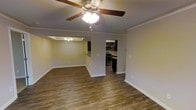 This single-story space showcases an open-concept layout that seamlessly connects the living area, dining area, and kitchen. The flooring throughout consists of rich, dark hardwood-style planks that add warmth and visual continuity across the space. Crown molding runs along the upper perimeter of the walls, lending a refined, finished appearance to the interior.

The living area is equipped with a ceiling fan featuring an integrated light fixture, providing both air circulation and ambient lighting. Adjacent to the living space, a defined dining area is distinguished by a tray ceiling accented with a small chandelier, adding an architectural focal point and a touch of elegance.

The kitchen is visible through an open passageway and features stainless steel appliances, including a range and an over-the-range microwave, alongside dark cabinetry. A peninsula or breakfast bar creates a natural transition between the kitchen and the dining area, enhancing the open flow of the layout.

The walls are painted in a neutral, light tone that complements the darker flooring, creating a balanced and versatile aesthetic throughout the space.
