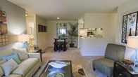This single-floor residence offers an open and inviting layout that seamlessly connects the main living spaces. The living room is generously sized, featuring plush wall-to-wall carpeting that adds warmth and comfort underfoot. Recessed lighting in the ceiling provides a clean, modern touch, while neutral-toned walls create a bright and versatile backdrop throughout the space.

The dining area sits centrally within the open floor plan, accommodating a round dining table and chairs, making it ideal for casual meals or entertaining. A tall indoor plant adds a refreshing natural element, enhancing the welcoming ambiance of the room.

Adjacent to the dining area, the kitchen is accessible via a pass-through counter, offering a practical and social connection between cooking and living spaces. The kitchen features white cabinetry and appliances, contributing to a clean and organized aesthetic. Upper cabinets provide ample storage, keeping the space tidy and efficient.

A hallway visible from the living room suggests additional rooms beyond, including what appears to be a bathroom or utility area. Throughout the space, the cohesive color palette of soft whites, beiges, and greens ties each area together, resulting in a harmonious and comfortable single-level home.