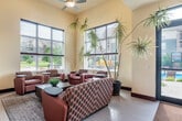 This expansive single-floor clubhouse offers a well-appointed communal environment spanning a generously sized open floor plan. The space is defined by soaring ceilings, warm neutral walls accented by bold deep-red architectural panels, and light hardwood-style flooring that flows seamlessly throughout.

The main living area serves as the heart of the space, anchored by a large stone-surround fireplace topped with a wall-mounted flat-screen TV. A substantial sectional sofa and decorative area rug create an inviting lounge setting. Tall windows on multiple walls flood the space with natural light and offer views of the surrounding outdoor areas, including a pool. A ceiling fan overhead adds to the airy atmosphere.

The fully equipped kitchen features a large central island with a stainless steel range hood, dark cabinetry with glass-front uppers, granite countertops, and a stone tile backsplash. A dedicated coffee station with rich espresso cabinetry provides a convenient self-serve beverage area.

A dining zone with bar-height seating and a billiards table contribute to the recreational character of the space. A separate work and dining nook with wall-mounted TV and workstation desks supports productivity. The welcoming entryway, highlighted by a recessed red ceiling detail, connects all areas cohesively into a vibrant, multi-functional communal space.