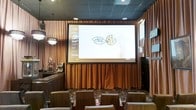 This entertainment venue is purpose-built for an immersive sports and media viewing experience. The centerpiece of the space is a large projection screen framed by warm brown floor-to-ceiling curtains, providing a cinematic atmosphere well-suited for live sporting events and broadcasts. The screen is complemented by a dark tiled ceiling fitted with recessed lighting, which maintains an ambient, low-lit environment ideal for viewing. A chandelier-style pendant light adds a touch of elegance to the otherwise casual setting. Lounge-style seating is arranged in a tiered or row-based layout facing the screen, offering guests clear sightlines from multiple vantage points. The patterned carpet flooring adds visual texture and contributes to the venue's relaxed, social atmosphere. Along one side of the room, a bar or service counter is visible, suggesting food and beverage service is available to guests. The combination of a dedicated screen wall, drapery accents, atmospheric lighting, and bar service makes this a well-equipped space for hosting sports watch parties, group screenings, and live event gatherings.