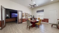 This single-story space presents a cohesive and well-appointed interior defined by a consistent design language throughout. A distinctive two-tone wall treatment — rich terracotta-hued wainscoting paired with crisp white upper walls — runs seamlessly across all rooms, lending a warm and unified aesthetic to the entire space.

The dining area is a standout, featuring soaring ceilings with a ceiling fan, recessed lighting, and generous natural light admitted through blinds-fitted windows with decorative fabric valances. Light wood-look flooring extends throughout, and tasteful wall art and a tall potted plant add character to the room.

Adjacent to the dining space is a private office, outfitted with a formal desk setup, ceiling fan, recessed lighting, and an open doorway that connects fluidly to the rest of the interior.

A lounge or sitting area provides a relaxed retreat, furnished with ample seating and framed artwork, and connects directly to the space's most dramatic feature: a dedicated home theater room. This darkened media room is equipped with a ceiling-mounted projector, a large projection screen, dual ceiling fans, dark curtain panels, and comfortable lounge seating — creating a true cinematic experience.

A central hallway with soaring ceilings and a reception-style pass-through window ties all areas together, completing this versatile and thoughtfully designed space.