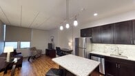 This thoughtfully designed single-floor space offers an open-concept layout that seamlessly blends the kitchen, dining, and living areas into one cohesive environment. The kitchen is a standout feature, equipped with stainless steel appliances including a full-size refrigerator and range, rich dark cabinetry, a light granite countertop, and a subtle tile backsplash that adds texture and visual interest. A granite-topped kitchen island serves as the centerpiece, illuminated by a sleek pendant track light fixture that lends a contemporary touch overhead. Recessed lighting complements the overall brightness of the space. The flooring throughout is warm hardwood, adding a sense of depth and elegance. Adjacent to the kitchen, a dedicated workspace features a desk and ergonomic chair, making the space functional for professional use. A cozy seating area with a patterned armchair and television rounds out the living space, creating a comfortable and versatile environment. The neutral wall tones and drop-tile ceiling contribute to a clean, modern aesthetic, while a window allows natural light to filter in, enhancing the open and welcoming ambiance of the space.