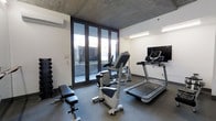 This dedicated fitness space combines modern design with functional amenities, offering an ideal environment for a comprehensive workout. The room is defined by its striking exposed concrete ceiling, which lends an industrial yet refined character to the space, complemented by crisp white walls and a durable dark-toned floor. A wall-mounted split-system air conditioning unit ensures a comfortable climate year-round.

Natural light pours in through a set of large floor-to-ceiling glass doors fitted with slim dark frames, which open directly onto an outdoor terrace or courtyard, providing a seamless connection to the exterior and a refreshing sense of openness. The glass frontage also allows for pleasant outdoor views during workouts.

The room is outfitted with a well-rounded selection of fitness equipment, including a treadmill, a stationary upright bike, and a weight bench — all arranged to maximize usability within the space. A dumbbell rack stocked with a graduated set of free weights offers additional strength-training options. A flat-screen television mounted on the wall provides entertainment and keeps the space engaging. Overhead strip lighting supplements the natural light, ensuring the room remains well-lit at all times. This fitness room delivers a complete, thoughtfully designed workout environment.