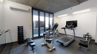 This dedicated fitness space combines modern design with functional amenities, offering an ideal environment for a comprehensive workout. The room is defined by its striking exposed concrete ceiling, which lends an industrial yet refined character to the space, complemented by crisp white walls and a durable dark-toned floor. A wall-mounted split-system air conditioning unit ensures a comfortable climate year-round.

Natural light pours in through a set of large floor-to-ceiling glass doors fitted with slim dark frames, which open directly onto an outdoor terrace or courtyard, providing a seamless connection to the exterior and a refreshing sense of openness. The glass frontage also allows for pleasant outdoor views during workouts.

The room is outfitted with a well-rounded selection of fitness equipment, including a treadmill, a stationary upright bike, and a weight bench — all arranged to maximize usability within the space. A dumbbell rack stocked with a graduated set of free weights offers additional strength-training options. A flat-screen television mounted on the wall provides entertainment and keeps the space engaging. Overhead strip lighting supplements the natural light, ensuring the room remains well-lit at all times. This fitness room delivers a complete, thoughtfully designed workout environment.