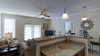 This single-floor space offers a well-designed open-concept layout that connects the kitchen, living area, and bedroom in a cohesive and functional arrangement. The kitchen is a standout feature, showcasing expansive granite countertops with rich, variegated patterning that extend into a peninsula or bar-style divider, complemented by warm wood cabinetry and a stylish pendant light fixture overhead. The open living area benefits from ample natural light streaming through double French-style doors dressed with horizontal blinds, creating a bright and airy atmosphere. A ceiling fan with a built-in light fixture serves both the aesthetic and comfort needs of the space. Crown molding adds a refined architectural detail along the upper walls, lending a polished feel throughout. The living area is furnished with a comfortable sofa and features a flat-screen television on a sleek metal stand, with coordinating side tables that maintain a consistent design sensibility. A glimpse through an open doorway reveals the bedroom beyond, which appears to continue the clean, neutral color palette established throughout the space. Overall, the layout and finishes create a cohesive, comfortable environment well-suited for everyday living.