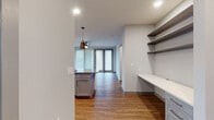 This single-floor residence showcases a thoughtfully designed open-concept layout with consistent wood-look flooring connecting the living spaces. The kitchen serves as a central highlight, featuring gray shaker-style cabinetry extending to the ceiling, white quartz countertops, a full suite of stainless steel appliances including a French door refrigerator, electric range, and microwave. A large center island with a contrasting gray countertop provides additional workspace and casual seating, illuminated by two modern pendant lights.

Adjacent to the kitchen, a dedicated built-in workspace features a long white desk surface and multiple open shelving units, ideal for a home office setup. The living area offers generous space with a ceiling fan, recessed lighting, and floor-to-ceiling glass doors that invite natural light.

The bedrooms are well-proportioned, each equipped with ceiling fans and large windows fitted with horizontal blinds. The primary bathroom features warm wood-toned cabinetry, a glass-enclosed shower, white tile flooring, and direct access to a spacious walk-in closet with built-in shelving. A secondary bathroom mirrors the clean aesthetic with subway-tiled tub surround and matching wood vanity.