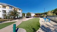 Located at 4005 Galantis Dr in Morehead City, North Carolina, this outdoor amenity space serves as a well-appointed dog park and recreational area within a multi-building residential community. The grounds are thoughtfully landscaped, combining a lush artificial turf putting green with generous gravel-covered open areas, providing a versatile environment for both pets and residents. A dedicated pet watering station is conveniently positioned within the space, ensuring comfort for four-legged visitors. Mature trees are scattered throughout, offering natural shade and a pleasant aesthetic against the backdrop of clear Carolina skies. The perimeter is fully enclosed by a sturdy black iron fence with gated access, providing a secure and contained environment. Concrete pathways and seating benches offer comfortable resting spots for residents. Decorative gravel landscaping borders the turf areas, minimizing maintenance while maintaining a clean, polished appearance. The surrounding multi-story residential buildings, finished in white and yellow siding, frame the space and contribute to its community-oriented character. Tall pine trees lining the outer fence add a natural, wooded backdrop. The overall ambiance is open, bright, and inviting, making it an ideal outdoor gathering and recreation space for residents and their pets.