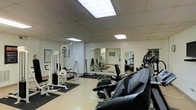 This fitness space is designed to accommodate a comprehensive workout experience within a single-floor layout. The room features a neutral color palette of white walls and a light-toned floor, creating a clean and open atmosphere. Recessed panel lighting in the drop ceiling provides consistent, even illumination throughout the space, ensuring excellent visibility for all activities.

The room is equipped with a variety of strength training machines, including a multi-function cable system and weight bench, positioned along one side of the space on protective rubber mats. A collection of free weights and dumbbells is neatly arranged on the floor nearby, offering additional versatility for resistance training. On the opposite side, several cardio machines, including what appears to be a treadmill and an elliptical trainer, are positioned for easy access.

Large wall-mounted mirrors span a significant portion of one wall, visually expanding the space and allowing users to monitor their form during exercise. Framed reference charts are displayed on the walls, adding a functional and informative element to the environment. Overall, the space is well-organized and purpose-built for effective physical training.