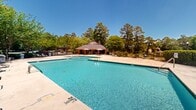 Located at Windsor Shores in Columbia, South Carolina, this outdoor community pool area is a well-appointed recreational amenity designed for relaxation and leisure. The centerpiece is a generously sized swimming pool with a distinctive L-shaped layout, offering both shallow and deeper sections clearly marked for swimmer safety. The crystal-clear, blue water is complemented by a broad concrete deck that provides ample space for lounge seating and poolside activities.

A charming covered pavilion with a hipped roof anchors one end of the pool area, offering shaded seating beneath open-air umbrellas — an ideal retreat from the South Carolina sun. The facility is enclosed by a chain-link fence with a gated entry, and signage at the entrance informs visitors of pool rules and the absence of a lifeguard on duty.

Surrounding the pool on all sides are mature trees and lush greenery, creating a naturally private and serene atmosphere. Additional amenities visible within the space include benches, waste receptacles, and a brick utility structure along the perimeter. The grounds are well-maintained, and the backdrop of tall pines and hardwoods enhances the outdoor ambiance, making this pool area a welcoming communal space within the Windsor Shores community.