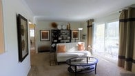 This single-story space centers around a thoughtfully designed living area that balances comfort and style. The living room is anchored by a tufted white sofa accented with warm orange throw pillows, complemented by a round glass-top coffee table that adds an open, airy feel to the room. Carpeted flooring runs throughout, contributing to a cozy and inviting atmosphere.

A dark wood bookcase serves as a prominent focal point along the back wall, flanked by two framed artworks that lend a curated, gallery-like quality to the space. Decorative accessories, including ceramic plates and sculptural accents, are displayed atop the shelving unit, adding visual interest and personality.

Natural light is a key feature, streaming in through a large sliding glass door dressed with floor-length tan curtains and dark accent panels. The door opens to an outdoor balcony or patio, offering views of surrounding greenery. Crown molding along the ceiling adds an architectural detail that elevates the overall finish of the space. A hallway visible in the background suggests additional rooms beyond the main living area.