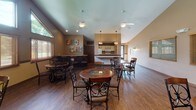 This inviting party room is designed to accommodate social gatherings with ease and comfort. The space is anchored by soaring vaulted ceilings that lend an open, airy feel, complemented by dual ceiling fans that keep the atmosphere comfortable. Warm wood-tone flooring runs throughout, tying the space together with a cohesive, welcoming aesthetic.

The main gathering area features multiple round dining tables with elegant wrought-iron and wood chairs, offering flexible seating arrangements for groups of various sizes. Soft wall sconces provide warm accent lighting throughout, while large windows dressed with wood blinds allow natural light to filter in, brightening the space.

A raised counter or bar area adds a social focal point, and framed artwork on the walls — including landscape prints and decorative signs — contribute a homey, personalized touch to the room's character.

A fully equipped galley-style kitchen is tucked conveniently off the main area, outfitted with a refrigerator, stove, dishwasher, double sink, and ample cabinetry, making it easy to serve food and beverages for events. Restroom access is also available from within the space.

The overall ambiance strikes a balance between casual warmth and functional versatility, making this party room a well-appointed venue for celebrations, community gatherings, or private events.