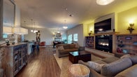 Located at 12925 East Mansfield Avenue in Spokane Valley, WA, this single-story community clubhouse offers a well-appointed gathering space designed for comfort and functionality. The welcoming entry area features elegant wingback chairs, a rustic wood accent table, and large glass doors that flood the space with natural light and provide views of the surrounding neighborhood.

The expansive main lounge serves as the heart of the facility, boasting vaulted ceilings, rich hardwood flooring, and a striking floor-to-ceiling fireplace flanked by built-in shelving. Comfortable seating arrangements and pendant lighting create a warm, inviting atmosphere. A dedicated dining area accommodates a large communal table with upholstered chairs, complemented by a decorative sideboard and patio door access.

The fully equipped kitchen features light maple cabinetry, dark granite countertops, black appliances, and a spacious bar-height island with industrial-style seating — ideal for communal gatherings. A convenient beverage station with upper cabinetry connects the common areas.

The fitness center is a standout feature, offering a generously sized workout space with rubber flooring, a full complement of strength training equipment, wall-mounted televisions, mirrored walls, and oversized garage-style windows that bring in abundant natural light. Additional multipurpose areas visible in the floorplan provide flexible space for various activities.