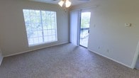 This single-floor space features a bright, open living area finished with wall-to-wall carpeting in a neutral speckled tone, creating a comfortable and understated foundation throughout. The room is filled with natural light from two notable sources: a large, horizontally oriented window fitted with horizontal blinds along one wall, and a sliding glass door dressed with sheer white curtains on the adjacent wall. The sliding door opens directly onto a wooden balcony, offering views of mature surrounding trees that lend a sense of privacy and a natural outdoor connection. A ceiling fan with an integrated light fixture is centrally mounted, providing both air circulation and ambient lighting. The walls are painted in a soft, neutral gray-beige tone, complementing the light carpeting and contributing to a clean, airy aesthetic. White baseboards frame the perimeter of the room, adding a crisp finishing detail. The overall layout feels open and functional, with ample floor space suitable for a variety of furniture arrangements. The combination of dual outdoor-facing openings and greenery views enhances the sense of openness and brings a refreshing connection to the outdoors into the living space.