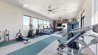 This fitness center offers a thoughtfully designed single-floor layout that provides ample space for a comprehensive workout experience. The main exercise area is bright and airy, benefiting from large windows along the perimeter that flood the space with natural light and offer views of the surrounding outdoor environment, including palm trees and adjacent amenities.

The flooring features a combination of tiled carpet and teal exercise matting, clearly delineating dedicated zones for cardio equipment and functional training. The cardio section is well-stocked with treadmills, elliptical trainers, stair climbers, and a stationary bike, all positioned to take advantage of the natural light and outdoor views. A large flat-screen television is mounted on the wall for entertainment during workouts.

The strength training area includes a selection of selectorized weight machines and a rack of kettlebells in varying weights, catering to a range of fitness levels. A striking large-format photographic mural depicting active individuals serves as a motivating focal point on one wall.

Overhead, large ceiling fans promote air circulation throughout the space. The facility also includes an adjoining restroom or changing area, along with a water station for convenience. The crisp white walls and modern finishes give the entire space a clean, contemporary aesthetic.