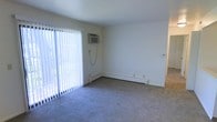 This single-floor space presents a straightforward and functional layout centered around a modestly sized living area. The room is finished with wall-to-wall carpeting in a neutral gray tone, contributing to a clean and understated aesthetic throughout. White walls and a white ceiling keep the space feeling bright and open.

A prominent sliding glass door fitted with vertical blinds serves as the primary source of natural light, filling the room with an airy quality while also providing access to what appears to be an outdoor area. Climate control is addressed through a combination of a wall-mounted air conditioning unit positioned above the sliding door and baseboard heating that runs along the lower perimeter of the room, offering year-round temperature management.

A doorway visible along the right side of the space suggests a corridor leading to additional rooms within the unit. The overall ambiance is clean and move-in ready, with a simple, no-frills design that provides a neutral canvas suitable for a variety of furnishing styles. The space is practical and functional, with all essential utility features conveniently integrated into the living area.