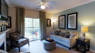 This single-floor residence offers a thoughtfully designed living space that blends comfort and style. The living room serves as the centerpiece of the home, featuring plush neutral-toned carpeting and a ceiling fan with integrated lighting that ensures year-round comfort. Crown molding adds a refined architectural detail, framing the room with a polished finish.

Natural light flows generously through a large sliding glass door, which opens directly onto a private balcony. The balcony overlooks a lush, tree-lined landscape, providing a serene outdoor retreat furnished with seating for al fresco relaxation. Floor-to-ceiling curtains with grommet detailing frame the glass door elegantly, offering both privacy and style.

The living area is furnished with a neutral upholstered sofa accented with decorative pillows, a tufted round ottoman, and a classic accent bench, creating an inviting and functional arrangement. A tall arc floor lamp provides warm ambient lighting, complementing the room's sophisticated atmosphere. The walls are adorned with a curated selection of framed botanical and decorative artwork, adding depth and character to the space.

Overall, the residence exudes a warm, welcoming ambiance with a tasteful blend of classic and contemporary design elements.