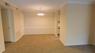 This single-story space showcases a well-proportioned main room finished with warm beige carpet and neutral-toned walls that create a clean, welcoming atmosphere. Crown molding lines the ceiling throughout, lending a touch of classic architectural detail, while a white chair rail runs along the perimeter walls, adding visual structure and a traditional design element. A multi-bulb chandelier hangs centrally from the ceiling, serving as a functional and decorative focal point that illuminates the room effectively. The layout features an open passageway on one side, connecting the main living or dining area to what appears to be a kitchen, promoting a natural flow between spaces. White baseboards and trim throughout provide crisp contrast against the neutral walls, contributing to a tidy, cohesive aesthetic. The space benefits from a straightforward, practical layout on a single level, making it accessible and easy to navigate. Overall, the interior presents a traditional, understated character with consistent finishes that provide a versatile foundation suitable for a variety of furnishing and decorating styles.