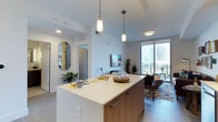 This contemporary single-floor residence showcases a thoughtfully designed open-concept layout that seamlessly connects the kitchen, dining, and living areas. The kitchen is a standout feature, anchored by a large island with a clean white quartz countertop and warm wood cabinetry, illuminated by a trio of stylish pendant lights that add both function and character. The space flows naturally into a casual dining area and a comfortable living room, all unified by large-format tile flooring that enhances the sense of openness. The living area is bathed in natural light streaming through expansive floor-to-ceiling sliding glass doors, which open to a balcony and frame striking views of a vibrant urban skyline. A flat-screen television is mounted on the accent wall, and curated décor including a decorative oval mirror, potted greenery, and a patterned area rug add warmth and personality throughout. The crisp white walls and recessed lighting contribute to a clean, airy ambiance, while the overall design strikes a balance between modern sophistication and everyday livability. The layout also features a private hallway leading to additional rooms, providing a clear separation between the living and private areas of the home.