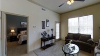 This 1-bedroom, 1-bathroom residence is designed to maximize comfort and functionality within a thoughtfully arranged single-floor layout. The living area immediately impresses with its soaring ceilings, which lend an expansive, airy quality to the space and enhance the sense of openness. Crown molding adds a refined architectural detail, while a ceiling fan provides both style and practical climate comfort. Natural light flows generously into the living room through large windows fitted with blinds, creating a warm and welcoming atmosphere. The neutral, light-toned walls offer a versatile backdrop that complements a variety of décor styles. Soft carpeting underfoot adds warmth and texture to the main living area. The bedroom, visible through an open doorway, continues the bright and neutral aesthetic, providing a restful retreat. The overall design balances simplicity with subtle elegance, making it an inviting and practical home. The layout connects the living and sleeping areas in a seamless flow, reflecting a well-considered use of space throughout the residence.