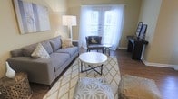 This single-floor space offers a comfortable and thoughtfully designed living environment with a warm, inviting ambiance throughout. The living area serves as the centerpiece of the space, showcasing rich wood-tone flooring that anchors the room with a sense of warmth and continuity. Neutral wall tones in soft beige provide a versatile backdrop that complements the contemporary furnishings, including a plush gray sofa, an accent armchair, and a round white coffee table atop layered geometric-patterned area rugs in gold and white tones. Natural light pours generously into the living area through elegant French doors dressed in sheer white curtains, which open to an outdoor area, creating a seamless connection between interior and exterior living. A stylish floor lamp adds additional ambient lighting, while carefully chosen décor pieces — including wall art and accent accessories — contribute to the space's polished yet relaxed aesthetic. The overall atmosphere is bright, airy, and modern, making it a highly functional and visually appealing space suited for everyday living and entertaining alike.