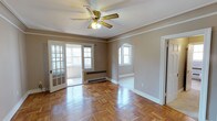 This single-floor space exudes classic character through a thoughtful combination of architectural details and practical features. The main living area showcases beautifully patterned parquet hardwood flooring that adds warmth and visual texture underfoot, complemented by crisp white crown molding that frames the ceiling with an elegant finish. A ceiling fan with a light fixture is centrally positioned, providing both air circulation and ambient lighting.

Natural light flows generously into the space through multiple windows, including a set of French doors that open to what appears to be a sunlit balcony or enclosed porch, enhancing the sense of openness. A charming arched doorway serves as a distinctive architectural element, connecting the main room to an adjacent space and adding a touch of vintage character. A baseboard radiator beneath one of the windows indicates a traditional heating system. The neutral warm-toned walls provide a versatile backdrop adaptable to a variety of interior styles. The overall ambiance is one of understated elegance, combining period-inspired details with a bright and welcoming atmosphere.