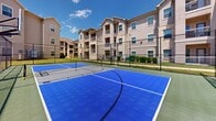 Located at 1101 Longhorn Rd in Fort Worth, Texas, this outdoor sport court is a well-appointed multi-use recreational facility. The court features a vibrant blue and gray interlocking tile surface with clearly marked lines for both basketball and tennis or pickleball play. A regulation-style basketball hoop with a clear backboard anchors the center of the court, while a removable net post system allows for versatile court configurations. The entire court area is enclosed by a sturdy black chain-link fence, providing both safety and defined boundaries for active play. The open-air design allows for abundant natural light and open sky views, creating an energetic outdoor atmosphere ideal for recreational use. Surrounding the court is a well-maintained grassy border, and the facility is set against the backdrop of a multi-story residential complex featuring a combination of stucco and stone exterior finishes with balconied units. The court's durable, weather-resistant surface and comprehensive sports markings make it suitable for a variety of athletic activities, serving as a central recreational amenity for the surrounding community in Fort Worth, TX.