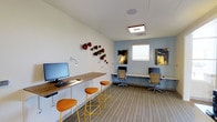 This single-floor workspace is thoughtfully designed to provide a functional and visually engaging environment for work and study. The room features multiple computer workstations, including a long wooden counter-height desk along one wall equipped with a desktop computer and paired with bright orange and yellow bar stools, adding a lively pop of color to the space. Along the opposite wall, a row of dedicated desk stations with ergonomic task chairs offers additional workspace, each position equipped with a monitor and ample surface area for individual use. Natural light filters in through windows positioned along the far wall, contributing to a well-lit and comfortable atmosphere. The interior walls are finished in a clean white and soft blue palette, creating a calm, focused ambiance. A distinctive wall art installation featuring sculptural red and gold circular elements serves as a creative focal point, lending personality and visual interest to the space. Patterned carpet flooring in neutral tones runs throughout, adding texture underfoot while contributing to a professional yet welcoming feel. Recessed and surface-mounted lighting fixtures ensure consistent illumination throughout the room, making this space well-suited for extended periods of productive work.