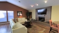 This single-floor space makes a strong first impression with its thoughtfully designed living area, where comfort and character come together seamlessly. The living room serves as the heart of the space, anchored by a classic brick fireplace that adds both warmth and timeless architectural charm. A large flat-screen television is mounted above the fireplace, making it a natural focal point for the room. Rich hardwood flooring runs throughout, complementing the earthy, warm color palette defined by deep terracotta-toned accent walls set against neutral cream tones. The living room is generously proportioned, offering ample space for a large sectional sofa and a round tiered coffee table, all set atop a decorative area rug with floral detailing. Recessed lighting and flush-mount ceiling fixtures provide even illumination throughout. Natural light filters in through a window, further brightening the interior. A decorative wall mirror and lush indoor plants add organic touches that enhance the inviting atmosphere. The layout flows naturally, with a visible transition to adjacent areas of the home. Overall, this space exudes a cozy, lived-in warmth that is both functional and aesthetically cohesive.