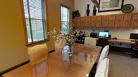This single-floor space exudes a warm and welcoming atmosphere, thoughtfully designed to balance everyday functionality with comfortable living. The dining area serves as a central highlight, featuring a gleaming glass-top table surrounded by elegantly upholstered chairs with classic nail-head trim detailing. A lush floral arrangement in a glass vase adds a fresh, organic touch to the polished surface. Natural light pours in generously through multiple windows dressed with wood-toned blinds, creating a bright and airy environment that complements the rich, warm tones found throughout the space. Adjacent to the dining area, a dedicated workspace or office nook is seamlessly integrated, complete with honey-toned wood cabinetry that extends to the upper walls for ample storage. Decorative black vases and framed artwork add refined accents to the built-in shelving above. The overall color palette draws from earthy tones — warm woods, creamy upholstery, and soft neutrals — lending the space a cohesive and inviting character. The layout flows naturally, making the space feel both organized and lived-in, ideal for those who value a blend of practicality and classic, understated elegance.