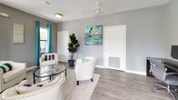 This single-floor space showcases a well-designed interior with a consistent and inviting aesthetic throughout. The main lounge area serves as the heart of the space, featuring light gray walls that provide a neutral backdrop for vibrant teal drapery panels that frame multiple windows, allowing generous natural light to fill the room. The flooring throughout is a smooth, light-toned wood-look surface that complements the overall palette. Comfortable cream upholstered armchairs are arranged around a circular glass-top coffee table atop a textured area rug, creating an approachable and welcoming seating arrangement. A wall-mounted mini-split climate control unit ensures year-round comfort. A pet-accessible door and tasteful abstract artwork add personality and practicality to the lounge. The entryway features classic double doors with glass panels that reinforce the flow of natural light into the interior. Adjacent to the lounge, a dedicated workspace area offers a long, dark-toned workstation desk accommodating two computer setups, each paired with ergonomic rolling chairs. A curated set of four framed abstract prints in coordinating teal and gray tones adorns the wall above the desk, maintaining the cohesive design theme. A decorative indoor plant adds a touch of greenery, enhancing the overall fresh and composed ambiance of the space.