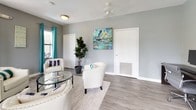 This single-floor space showcases a well-designed interior with a consistent and inviting aesthetic throughout. The main lounge area serves as the heart of the space, featuring light gray walls that provide a neutral backdrop for vibrant teal drapery panels that frame multiple windows, allowing generous natural light to fill the room. The flooring throughout is a smooth, light-toned wood-look surface that complements the overall palette. Comfortable cream upholstered armchairs are arranged around a circular glass-top coffee table atop a textured area rug, creating an approachable and welcoming seating arrangement. A wall-mounted mini-split climate control unit ensures year-round comfort. A pet-accessible door and tasteful abstract artwork add personality and practicality to the lounge. The entryway features classic double doors with glass panels that reinforce the flow of natural light into the interior. Adjacent to the lounge, a dedicated workspace area offers a long, dark-toned workstation desk accommodating two computer setups, each paired with ergonomic rolling chairs. A curated set of four framed abstract prints in coordinating teal and gray tones adorns the wall above the desk, maintaining the cohesive design theme. A decorative indoor plant adds a touch of greenery, enhancing the overall fresh and composed ambiance of the space.