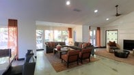 This single-floor space offers an open and inviting layout designed for comfort and social interaction. The lounge area serves as the centerpiece, featuring a well-coordinated seating arrangement that includes charcoal-toned sofas, rich burnt-orange accent chairs, and a sturdy wooden coffee table, all anchored by a warm, patterned area rug. Decorative throw pillows and a stylish table lamp add layers of warmth and texture to the setting.

Large floor-to-ceiling windows line the perimeter of the space, bathing the interior in abundant natural light while offering clear views of a lush outdoor area. Elegant burnt-orange drapery frames the windows, tying together the warm color palette that runs throughout the room. The polished concrete flooring extends across the open floor plan, lending an understated, contemporary feel to the overall aesthetic.

Recessed lighting in the white ceiling provides additional illumination, ensuring the space remains bright and functional at all hours. The seamless flow between seating zones and the generous sense of openness make this a versatile and welcoming environment suited for both relaxed gathering and everyday use.