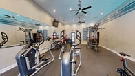 This fitness center offers a comprehensive workout environment designed to accommodate a wide range of exercise routines. The space is laid out on a single floor and makes efficient use of its generous footprint, providing distinct zones for cardio training, strength work, and functional fitness. A decorative teal stripe accent runs along the upper portion of the neutral-toned walls, lending the space a clean, modern aesthetic. Rubber flooring throughout ensures durability and safety during workouts. Recessed lighting and dual ceiling fans with integrated lights keep the space well-lit and ventilated. Large wall-mounted mirrors along one side visually expand the room while allowing users to monitor their form. The cardio area is equipped with treadmills, elliptical machines, and stationary and spin bikes, accompanied by wall-mounted flat-screen televisions for entertainment. A multi-station cable machine anchors the strength training zone, complemented by a dumbbell rack with a full range of free weights, a weight bench, and additional functional training equipment. Natural light filters in through windows overlooking the outdoor surroundings, and a water fountain is conveniently located near the exit door. Posted fitness center rules on the walls reinforce a structured, community-oriented atmosphere.