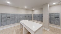 The Mail Center is a dedicated, single-floor facility designed to streamline mail and package management for residents. The space is thoughtfully laid out, with banks of gray metal mailboxes lining multiple walls, providing a large number of individual units with secure key-lock access. The mailboxes are organized in multi-row configurations, maximizing capacity while maintaining a clean, orderly appearance throughout the room.

At the center of the space sits a white countertop surface, offering a convenient area for sorting and organizing mail upon retrieval. The room features light wood-look flooring that complements the neutral beige walls, contributing to a bright and welcoming atmosphere. Recessed ceiling lighting ensures the space is well-lit throughout.

A glass-paneled door connects the mail center to an adjacent corridor, allowing for easy access from the building's common areas. Safety features, including a mounted fire extinguisher, are visibly present. The overall design prioritizes functionality and accessibility, with an open, uncluttered layout that allows for comfortable movement between the mailbox stations.