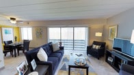 This single-floor space offers a comfortable and well-appointed living environment designed with modern sensibilities. The living area serves as the heart of the space, anchored by large sliding glass doors fitted with vertical blinds that flood the room with natural light while providing access to an outdoor balcony area. The expansive windows frame views of the surrounding greenery, adding a refreshing connection to the outdoors.

The living room is tastefully arranged with a deep navy blue tufted sofa accented by decorative throw pillows, complemented by a matching armchair that creates an inviting conversational seating area. A geometric-patterned area rug in shades of gray, yellow, and white grounds the space, adding a vibrant yet cohesive visual element underfoot. A dark-toned coffee table anchors the seating arrangement and provides functional surface space.

The ceiling features a distinctive shiplap detail that adds architectural character and a subtle texture to the room. Neutral wall tones provide a clean backdrop, enhanced by a colorful piece of wall art that introduces a pop of personality. A floor lamp positioned near the armchair ensures warm, ambient lighting for evenings. The overall ambiance is bright, modern, and inviting, striking a balance between comfort and contemporary style.