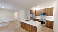 This thoughtfully designed 2-bedroom, 2-bathroom residence in Greenwich, CT offers a refined modern aesthetic throughout its well-appointed single floor. The open-concept living and kitchen area serves as the heart of the home, showcasing striking herringbone-pattern hardwood flooring that flows seamlessly through the space. The chef's kitchen is a standout feature, equipped with sleek walnut cabinetry, a large waterfall-edge quartz island with an undermount sink and brushed gold faucet, stainless steel appliances including a gas range, built-in microwave, and French door refrigerator, all illuminated by track lighting above.

Both bedrooms are bright and serene, each featuring black-framed grid windows that frame lush green outdoor views and continue the herringbone hardwood flooring. The generously sized primary bedroom includes access to well-organized walk-in closets outfitted with custom shelving, hanging rods, and gold hardware. Additional closets provide ample storage throughout.

The two bathrooms are elegantly finished with large-format tile flooring, square subway-tiled walls, floating walnut vanities with white quartz countertops, and coordinating brushed gold fixtures and mirrors. One bathroom features a full soaking tub with shower. A dedicated laundry room and utility room round out this complete and well-equipped residence.