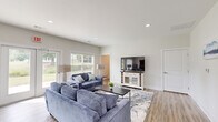 Located in Selmer, TN, this single-floor clubhouse offers a welcoming, open-concept layout that seamlessly combines a living room and dining area in one well-appointed space. The interior is finished with light-toned luxury vinyl plank flooring throughout, complementing the crisp white walls and creating a clean, modern aesthetic.

The living area is bright and generously proportioned, anchored by comfortable blue upholstered sofas arranged around a dark coffee table atop a patterned area rug. A white media console with glass-front cabinetry houses a flat-screen television, serving as a functional focal point for the room. Recessed ceiling lighting provides consistent illumination throughout.

Large windows fitted with horizontal blinds line the walls, bathing the space in natural light while offering views of the surrounding greenery. A set of French double doors provides direct access to the outdoors, enhancing the open, airy feel of the room.

The dining area is a distinct yet connected space, featuring a classic wood dining table paired with white ladder-back chairs. Subtle wall art adds a decorative touch without overwhelming the understated, neutral palette. The dining space flows naturally toward an adjacent kitchen area, making the clubhouse well-suited for gatherings and community events. Overall, the space conveys a fresh, inviting atmosphere ideal for shared use.
