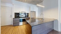 Located at 205 Rue De l'Équinox in Brossard, Quebec, this modern 1-bedroom, 1-bathroom unit showcases a thoughtfully designed open-concept layout finished with contemporary details throughout. The kitchen is a standout feature, boasting stainless steel appliances including a built-in oven, full-size refrigerator, and dishwasher, complemented by sleek dark cabinetry, a bold tile backsplash, tall white upper cabinets, and a large island with a stone countertop and integrated sink. Exposed concrete ceilings run throughout the main living area, lending an industrial-chic character that contrasts beautifully with the warm wood-look flooring and crisp white walls. A wall-mounted mini-split system ensures year-round comfort. The bedroom offers a calm, minimalist retreat with a window framing urban views and a baseboard heating unit. Adjacent to the bedroom is a dedicated walk-in closet fitted with wire shelving and hanging rods for ample storage. The bathroom is finished with neutral tile surrounding a full soaker tub and shower, paired with a modern floating vanity, integrated sink, and a large mirror. The unit's efficient, well-organized floor plan makes excellent use of every square meter, delivering a functional and stylish living space.