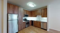 This single-floor space is designed with a consistent modern aesthetic, highlighted by warm walnut-toned cabinetry and light hardwood flooring throughout the main living and kitchen areas. The kitchen is a standout feature, outfitted with a full suite of stainless steel appliances including a gas range with overhead microwave, a full-size refrigerator, and a dishwasher. Ample upper and lower cabinetry with sleek bar-pull hardware provides generous storage, while white tile backsplash and light countertops create a clean, contemporary contrast to the rich wood tones. Under-cabinet lighting adds both functionality and ambiance. A secondary area replicates the kitchen's design language with matching cabinetry, a double stainless steel sink, a wine cooler, and a dishwasher, functioning effectively as a wet bar or prep station. The living area features neutral carpet and clean white walls, with a ceiling fan and a recessed upper ledge adding architectural interest. The bathroom is finished with a wood-framed vanity mirror, a white countertop with an integrated sink, matching walnut cabinetry, and a full soaking tub with floor-to-ceiling white tile surround, completing the space with a clean, cohesive finish.