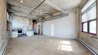 This single-floor loft showcases a striking industrial aesthetic defined by its exposed concrete ceiling with raw wood plank impressions, visible structural beams, and prominent silver ductwork running the length of the space. The open-concept layout creates a fluid connection between the spacious carpeted living area and the kitchen, which features a peninsula-style breakfast bar, pendant lighting, white cabinetry, and standard appliances. A ceiling fan centered in the main living area aids in air circulation while complementing the industrial design. The concrete flooring transitions to carpet in the living zone, offering a subtle contrast in texture and comfort. Natural light filters in, casting warm patches of sunlight across the floor and highlighting the interplay of raw and refined materials throughout. The neutral white walls provide a clean backdrop against the rugged ceiling elements, creating a balanced aesthetic that is both modern and utilitarian. Additional doors visible along the wall suggest access to private rooms or utility spaces, rounding out the functional layout. Overall, the space delivers an authentic urban loft experience with a blend of industrial character and practical, everyday livability.
