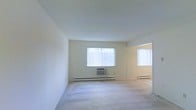 This single-floor space presents a clean and functional layout defined by its neutral color palette and practical features. The main living area is finished with wall-to-wall carpet flooring, offering a soft underfoot feel throughout. Crisp white walls and a smooth ceiling contribute to a bright, airy atmosphere. Natural light enters generously through multiple windows dressed with vertical blinds, allowing for adjustable light control while maintaining privacy. A wall-mounted air conditioning unit is positioned beneath one of the windows, complementing the baseboard heating system that runs along the perimeter of the room — together providing year-round climate comfort. An open doorway visible toward the far end of the space suggests additional rooms beyond, hinting at a functional floor plan. The overall ambiance is minimal and move-in ready, with a neutral foundation that lends itself well to a variety of interior styles. The space is well-lit, uncluttered, and benefits from a straightforward design that prioritizes utility and comfort in equal measure.